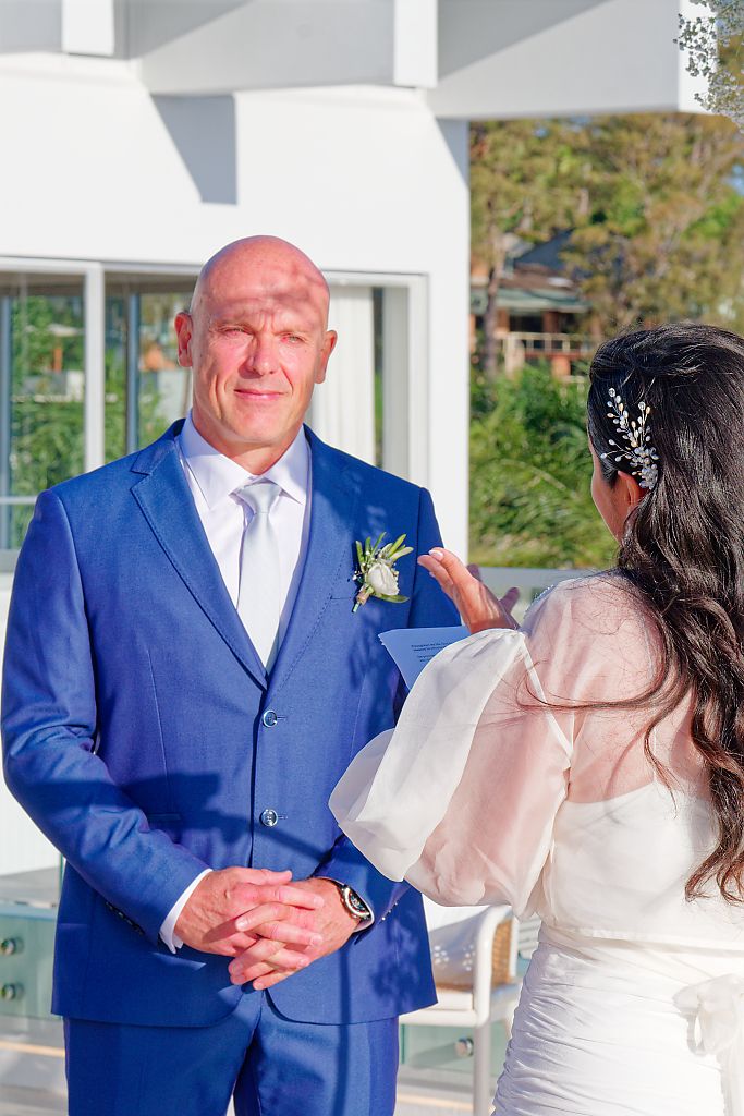 Groom in a blue suit with a white boutonniere clasps his hands and smiles during an outdoor wedding ceremony as the bride in white gestures.