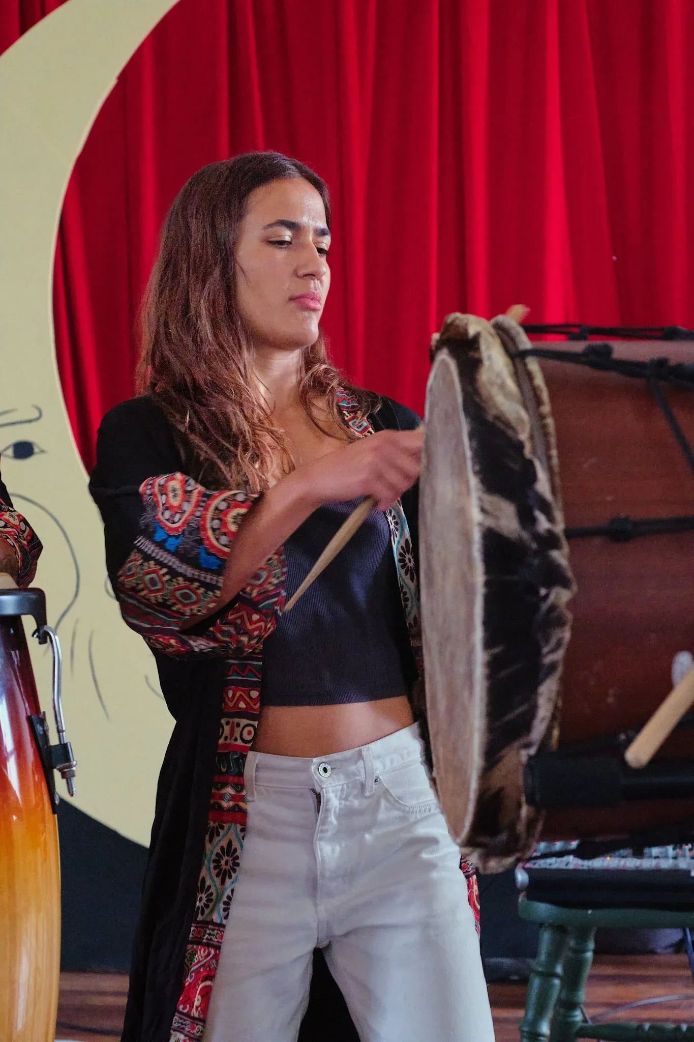 Female drummer plays a hand drum with a stick in front of red curtains and a crescent moon.
