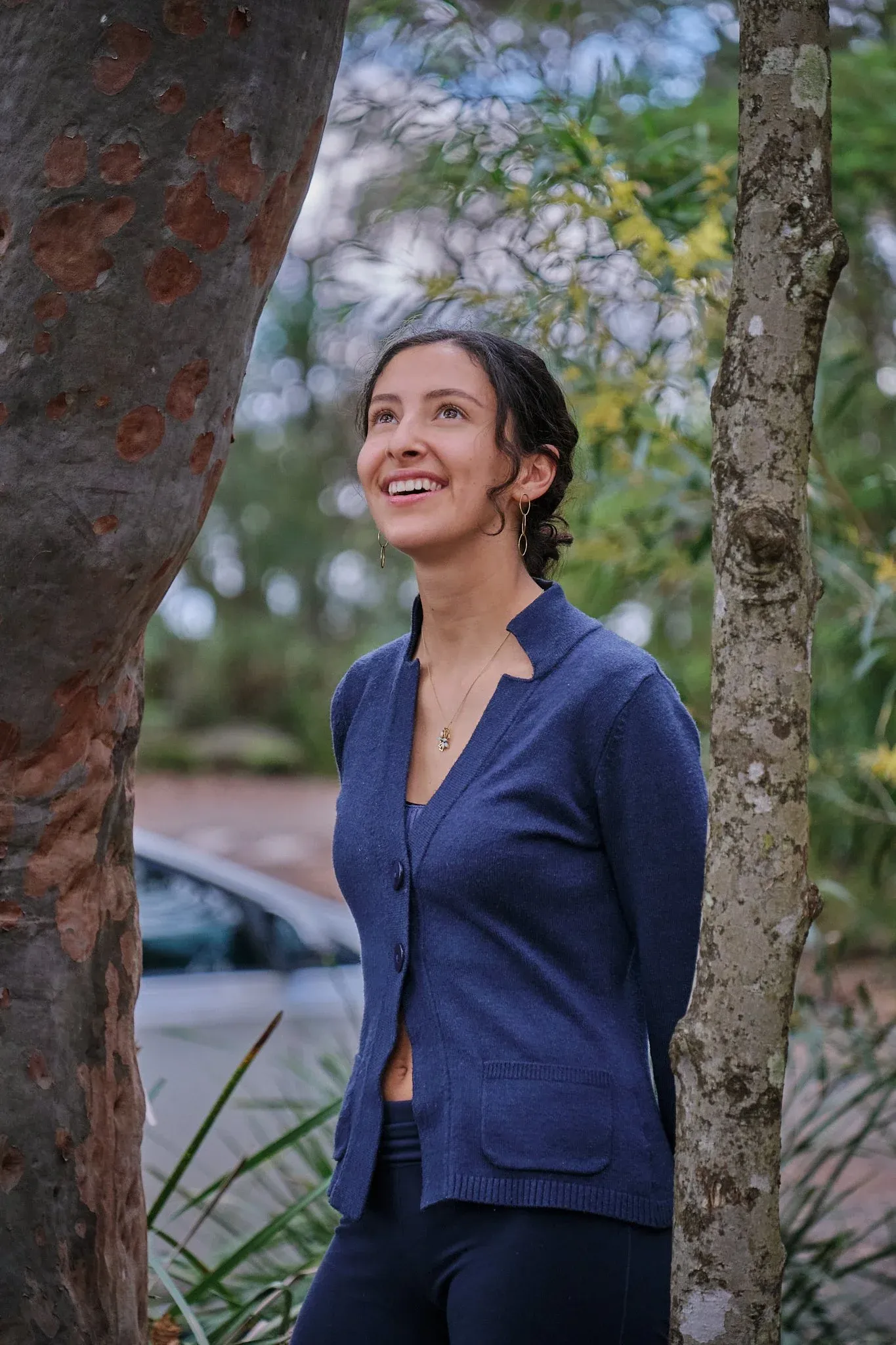Smiling woman in navy blue cardigan standing between trees in lush garden.