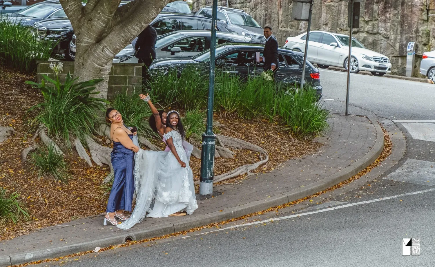 Bride and bridesmaid posing playfully near tree on curved street.