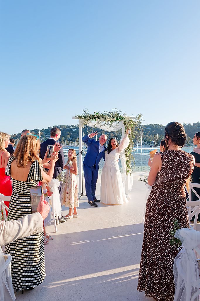 Newlyweds raise their arms in celebration under a floral arch as guests cheer and take photos at a waterfront wedding ceremony.