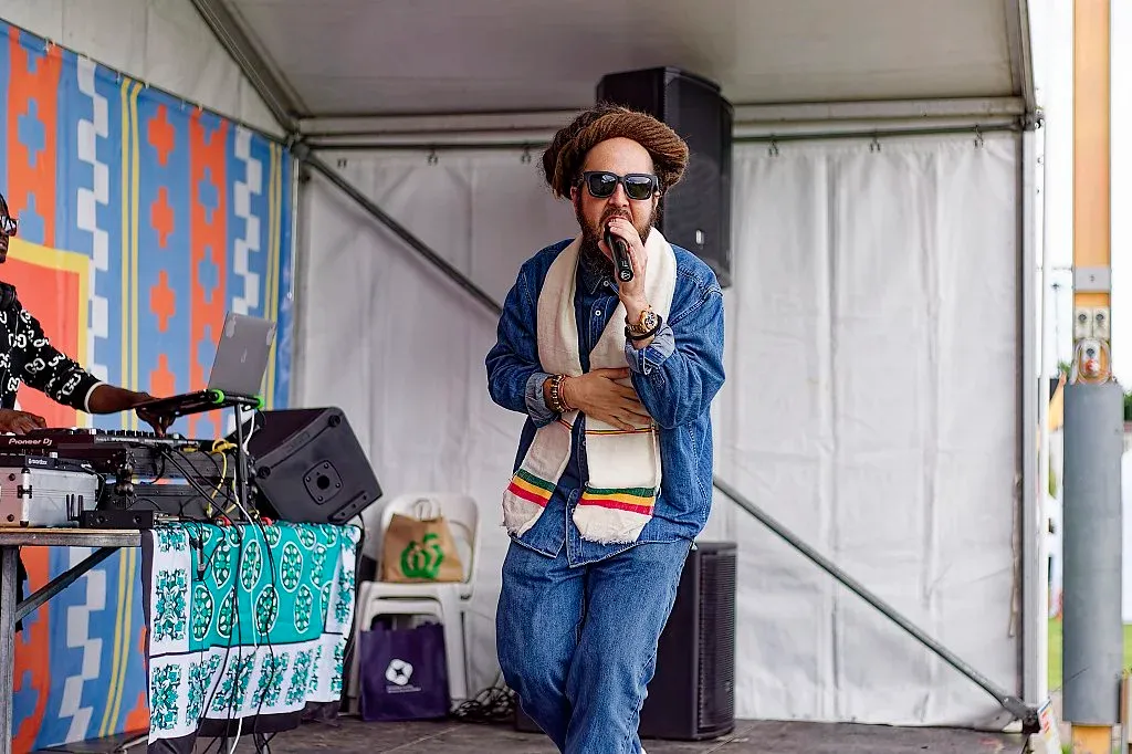 Performer in denim outfit and hat singing into microphone on colorful outdoor festival stage.