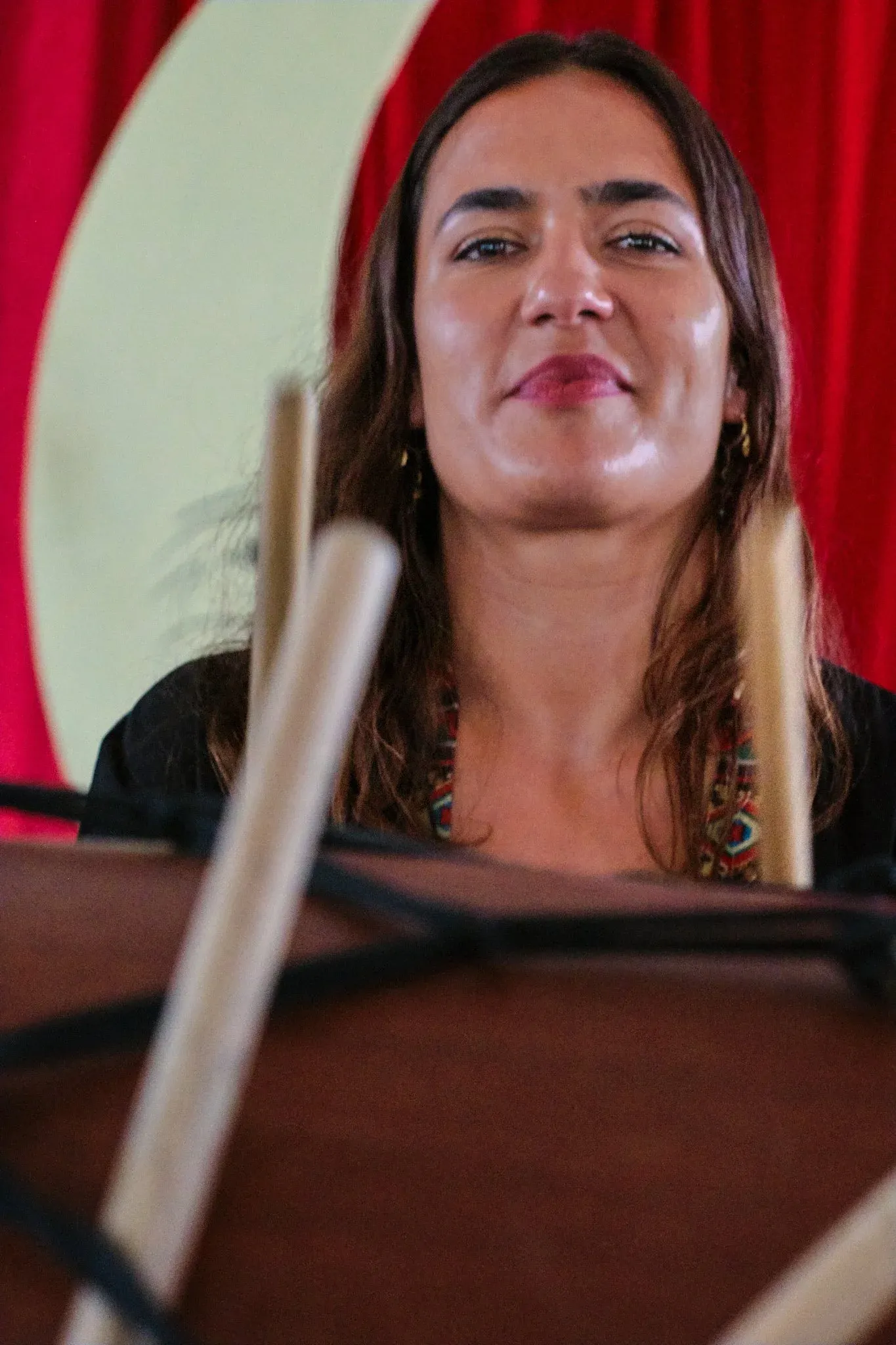 Woman musician performing behind a music stand with drumsticks in foreground, red curtain backdrop.