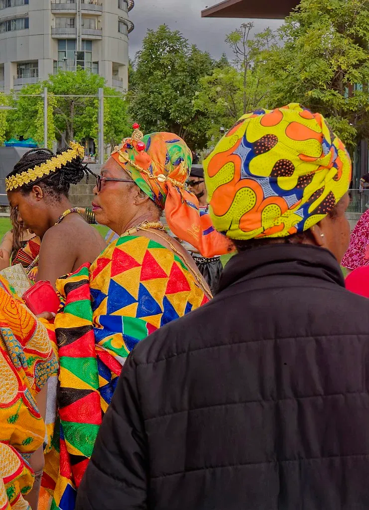 Women in colorful African kente cloth outfits and head wraps at outdoor cultural event.