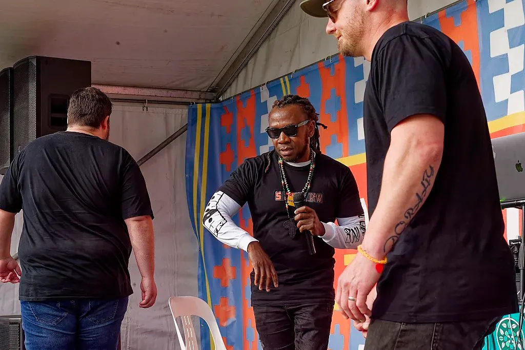 Performer in black and gold outfit interacting with crew members backstage at outdoor festival.