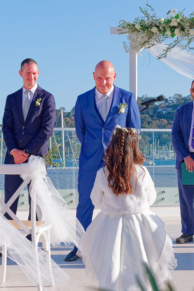 Two grooms in blue suits smile at a waterfront wedding ceremony beneath a floral arch while a flower girl in white looks on.