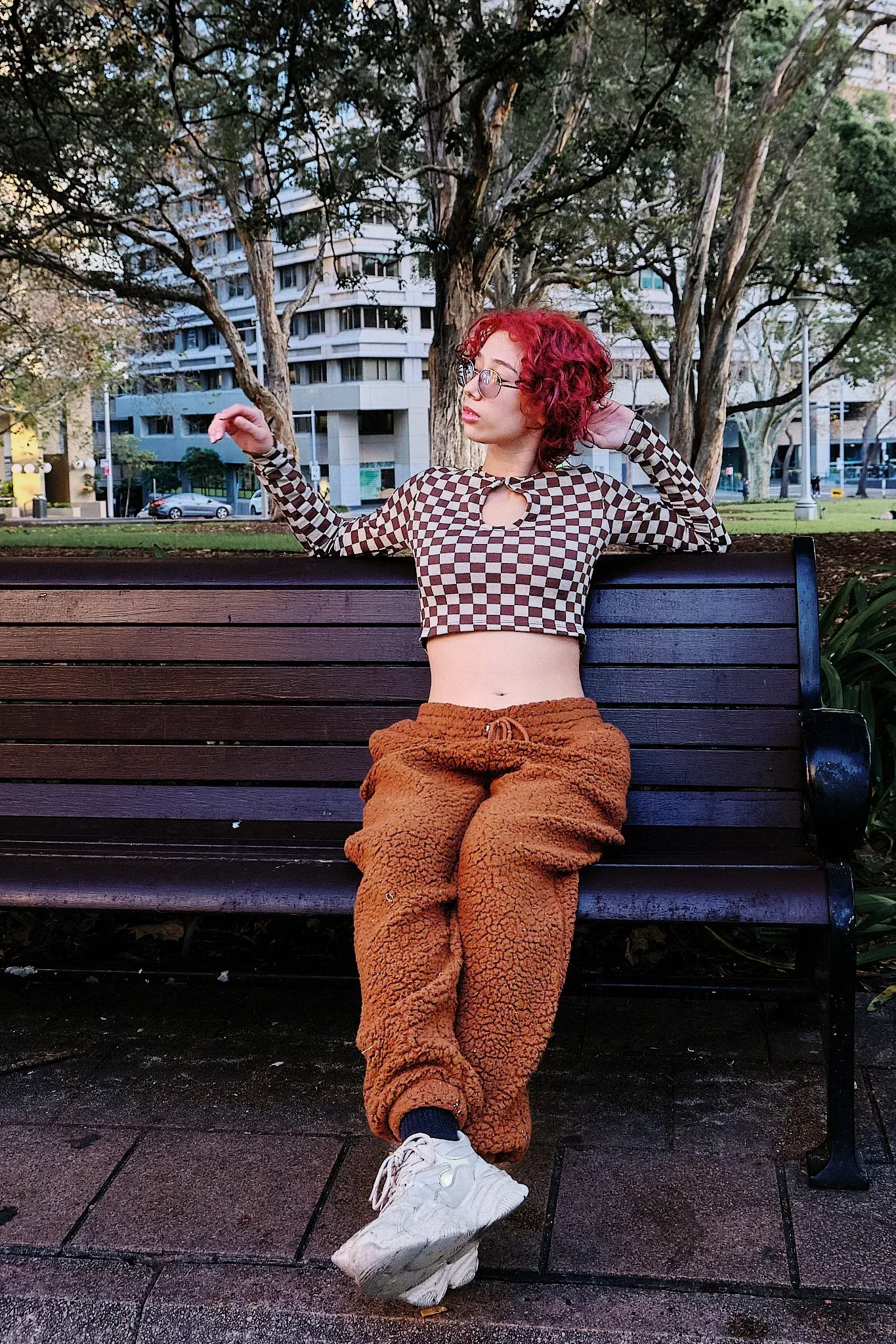 Red-haired woman in checkered crop top and brown fluffy pants sitting on park bench.