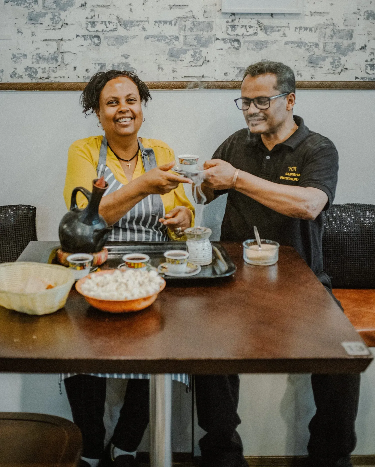 Two people sharing an Ethiopian coffee ceremony, clinking small traditional cups at a restaurant table.