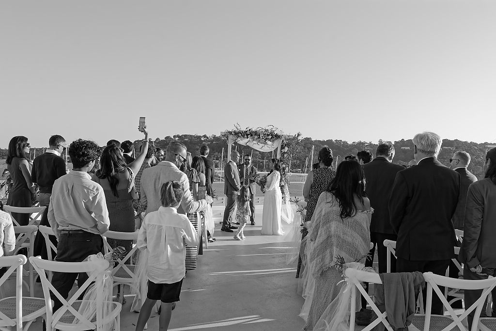 Wedding guests seated in white chairs watch a bride in a white gown walk toward a floral arch on a sunlit waterfront terrace.