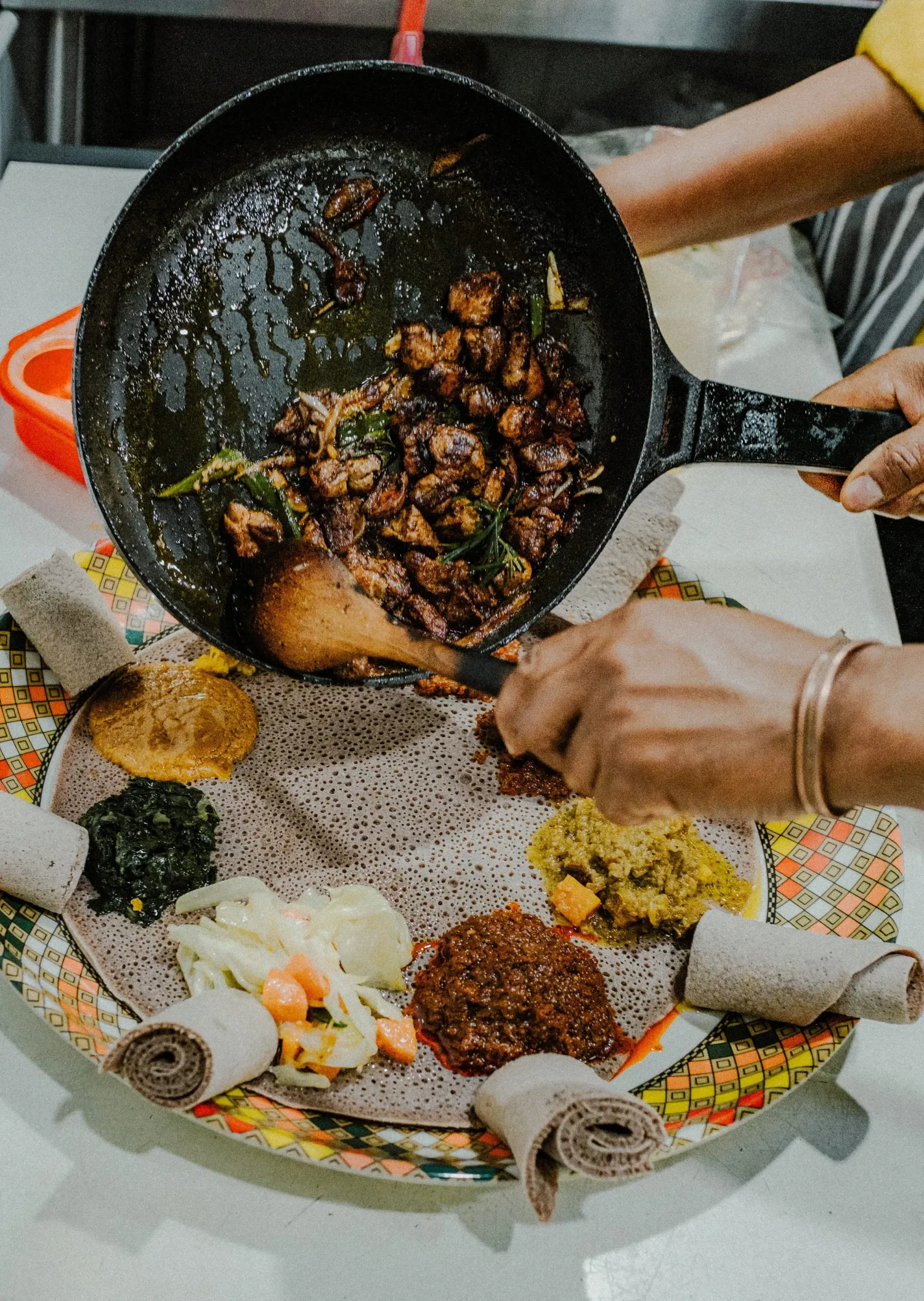Hands serving tibs from a cast iron skillet onto a colorful Ethiopian injera platter with various stews.
