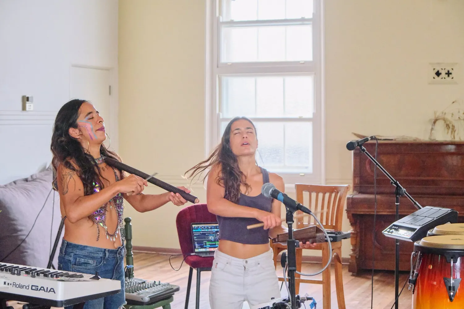 Two women laugh and sing together into a microphone during an indoor music rehearsal session.