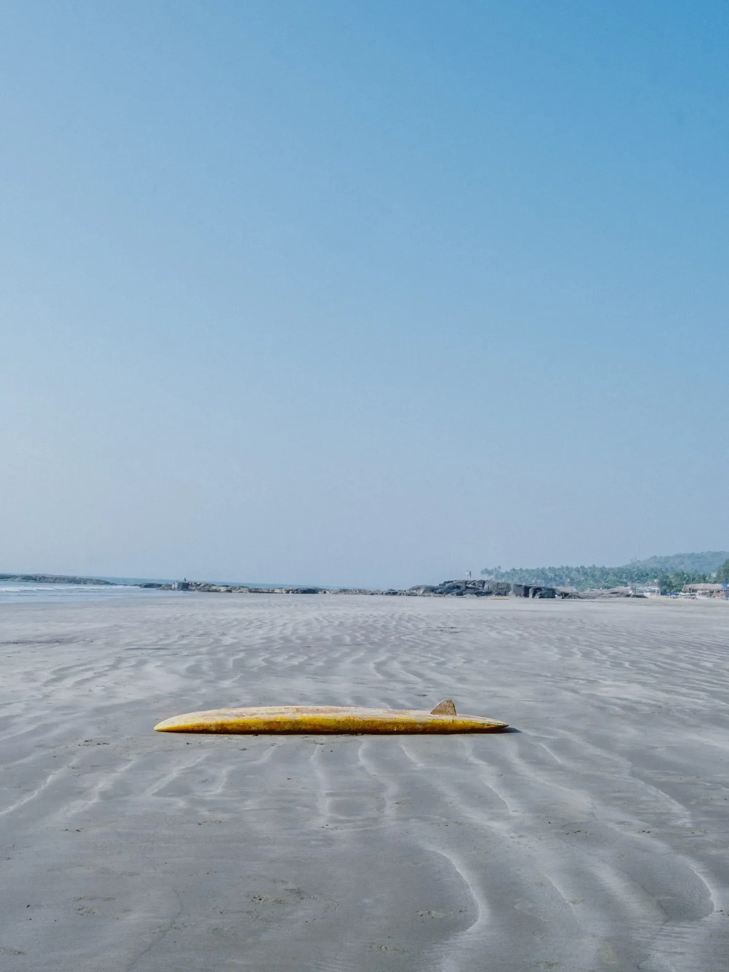 Yellow surfboard lying on a vast sandy beach under a clear blue sky.