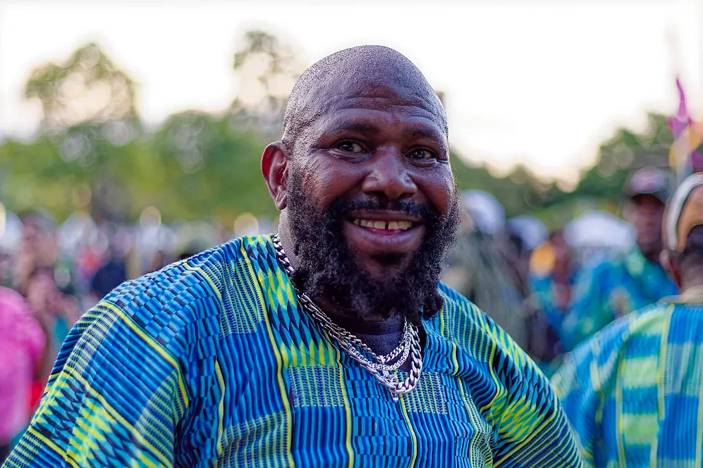 Smiling man with beard wearing vibrant blue and green African print shirt at outdoor cultural festival.