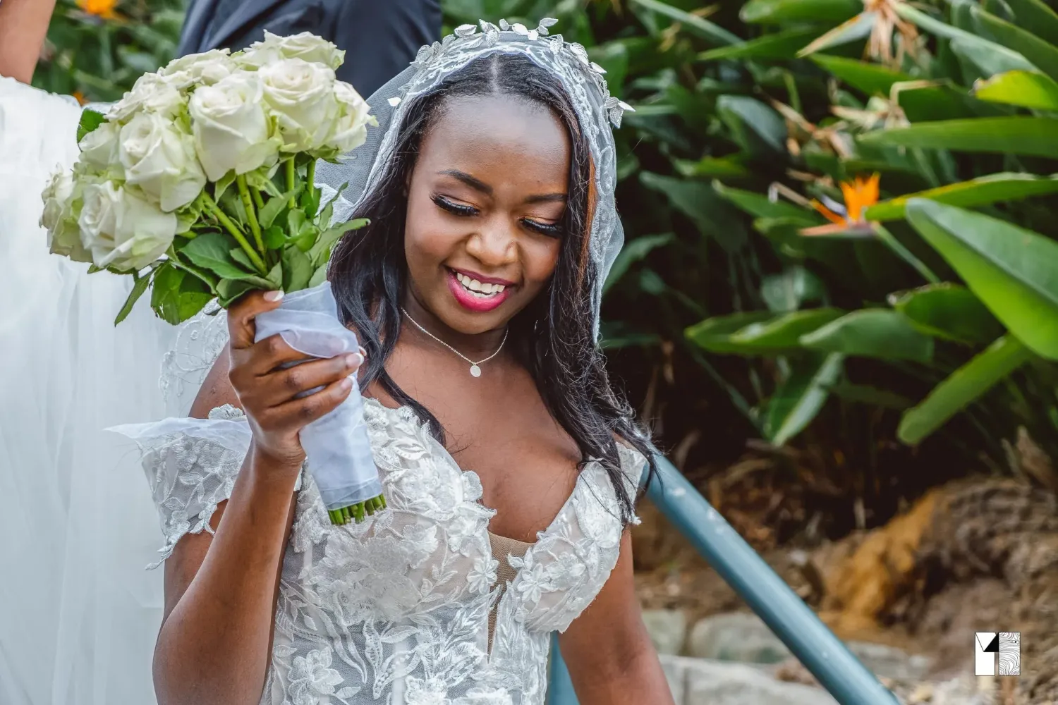Smiling bride in lace gown holding white rose bouquet in tropical garden setting.