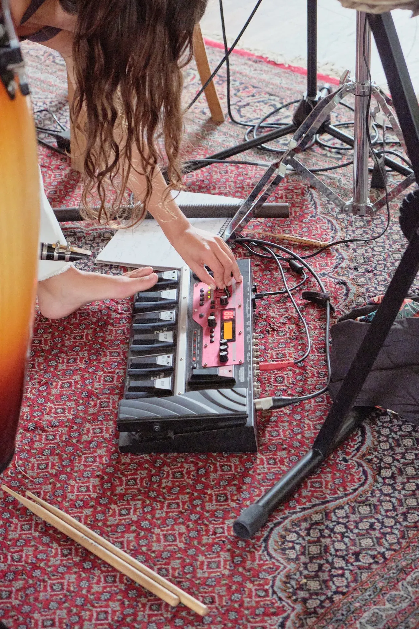 Musician adjusting a pink electronic bass pedal keyboard on a patterned rug with cables and drumsticks.