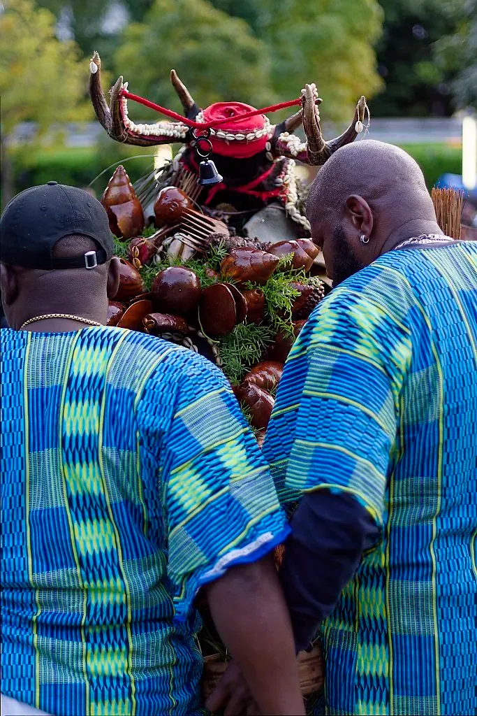 Two men in blue kente shirts viewing elaborate traditional African ceremonial mask display.