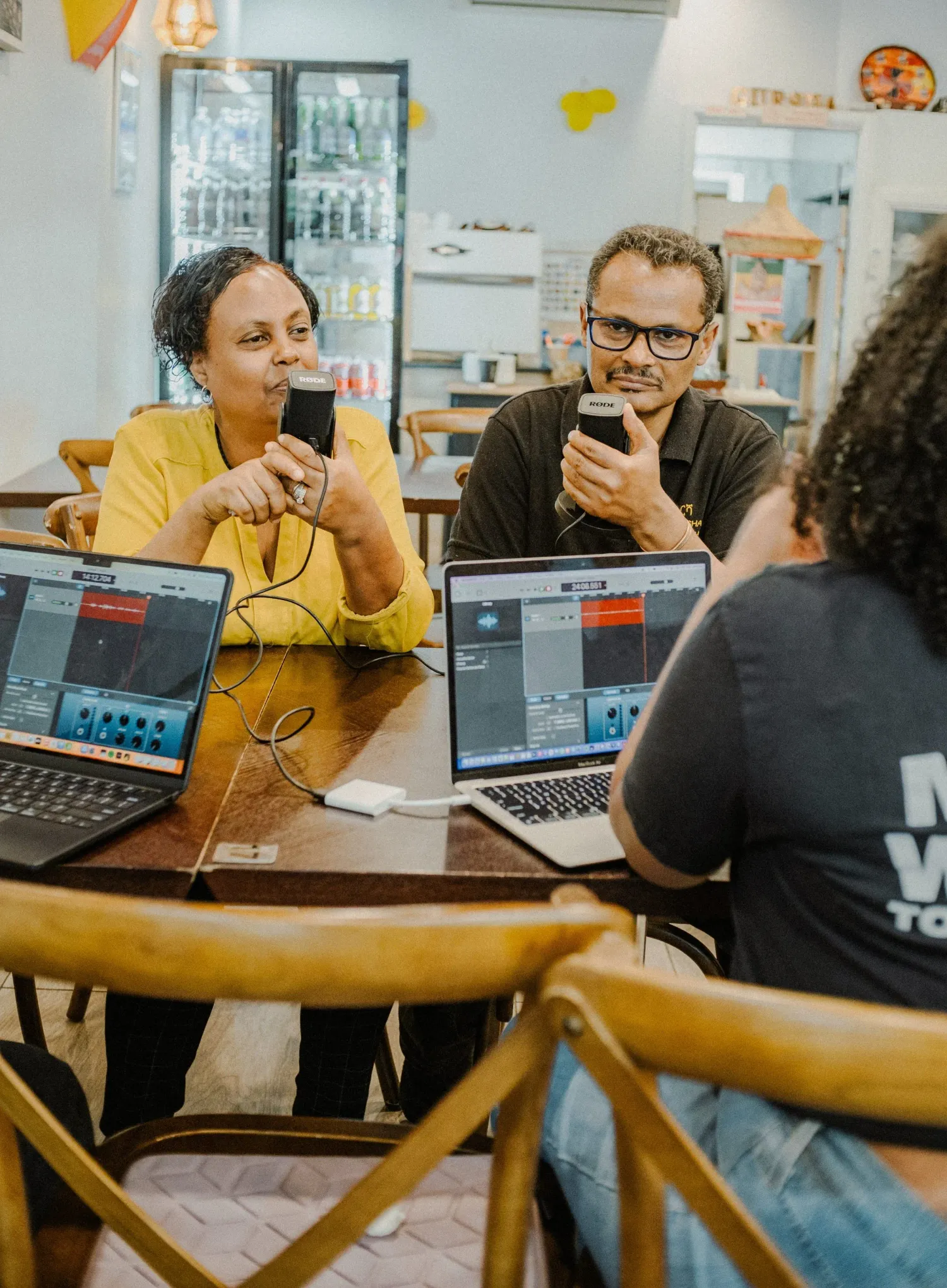 Three people recording a podcast with laptops and microphones at a cafe table.