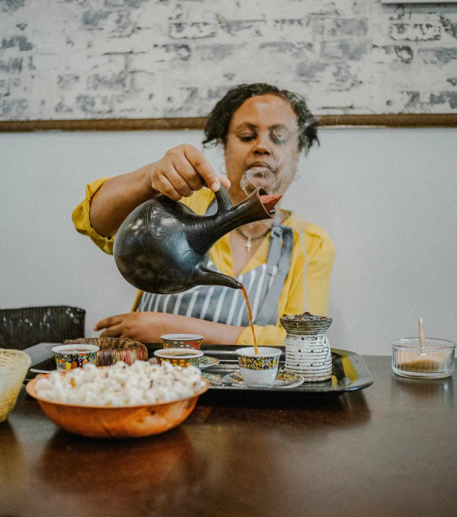 Woman in yellow shirt performing traditional Ethiopian coffee ceremony, pouring from a black jebena pot.