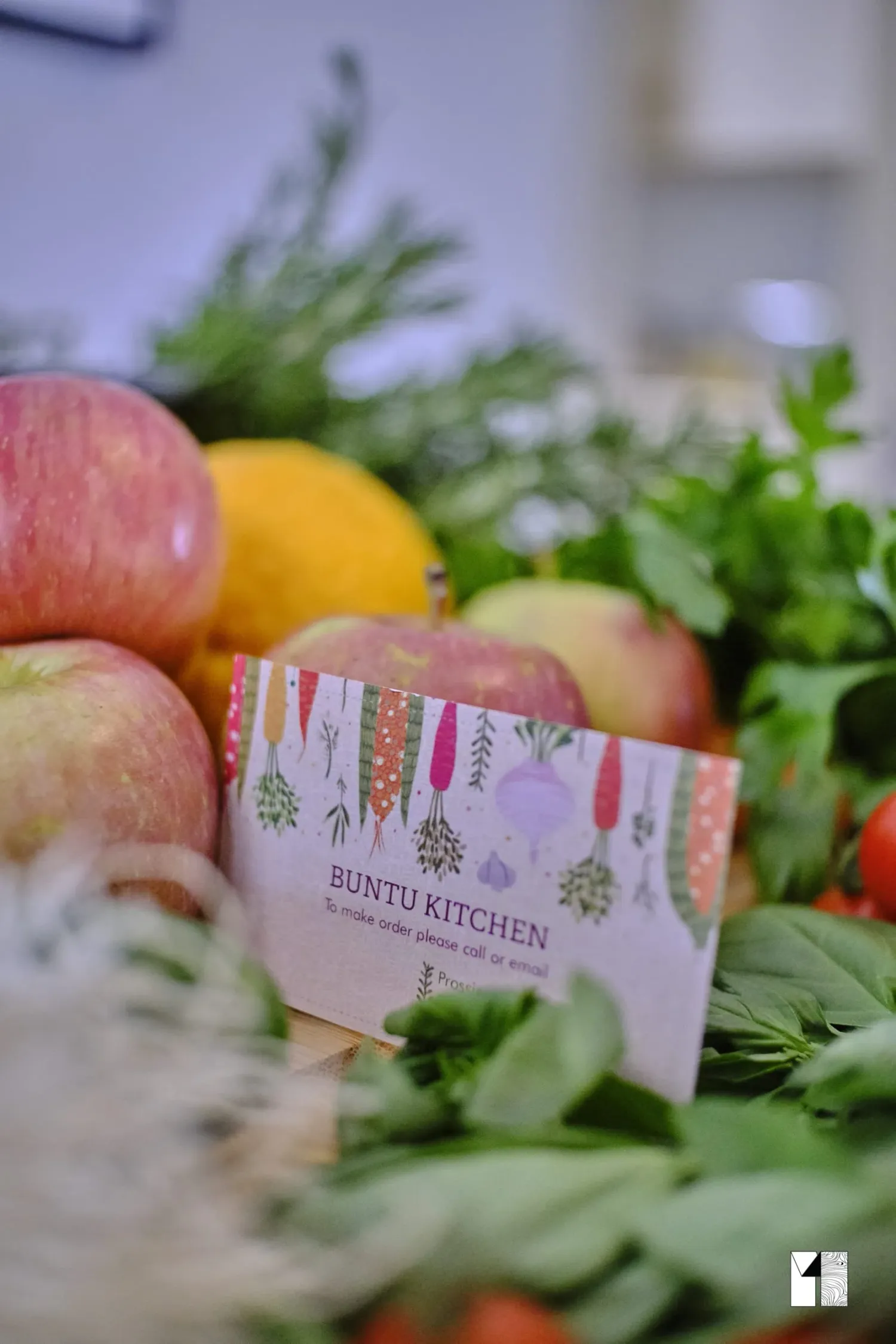 Buntu Kitchen business card surrounded by fresh apples, herbs, and colorful vegetables.