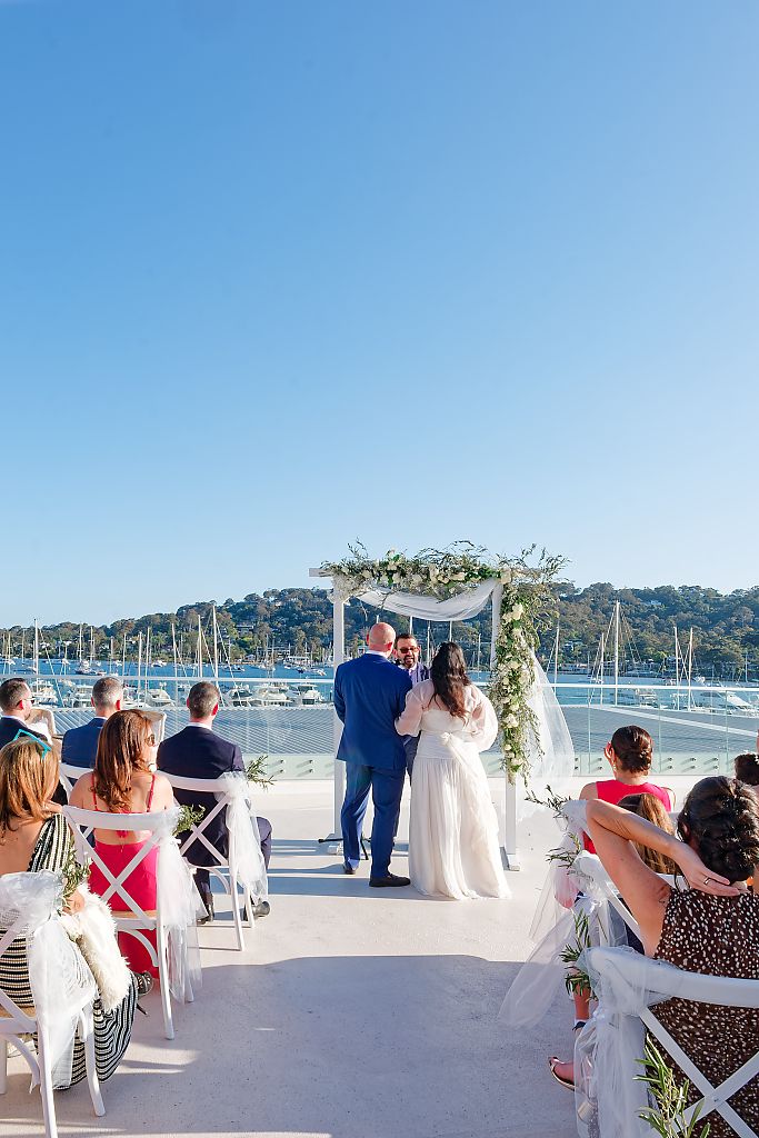 Bride and groom stand under a floral arch during an outdoor waterfront wedding ceremony as guests watch with a marina and sailboats behind.