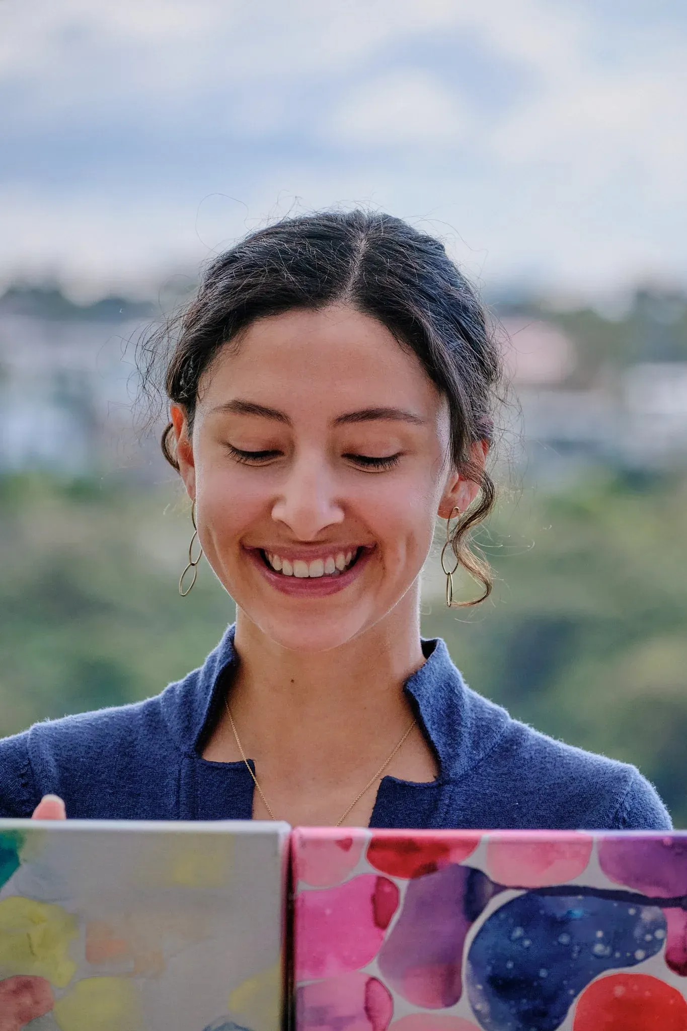 Smiling woman in blue top looking down at colorful abstract paintings with sky background.