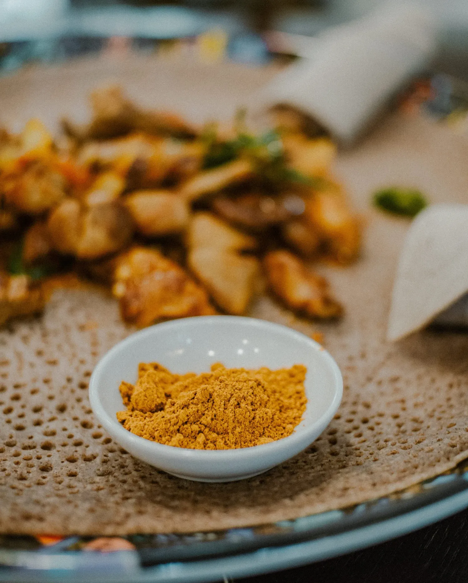 Small bowl of golden spice powder on injera beside seasoned chicken pieces on an Ethiopian platter.