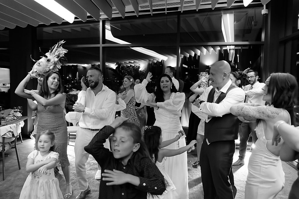 Black and white photo of wedding guests, including smiling children, dancing and cheering on a lively reception dance floor.