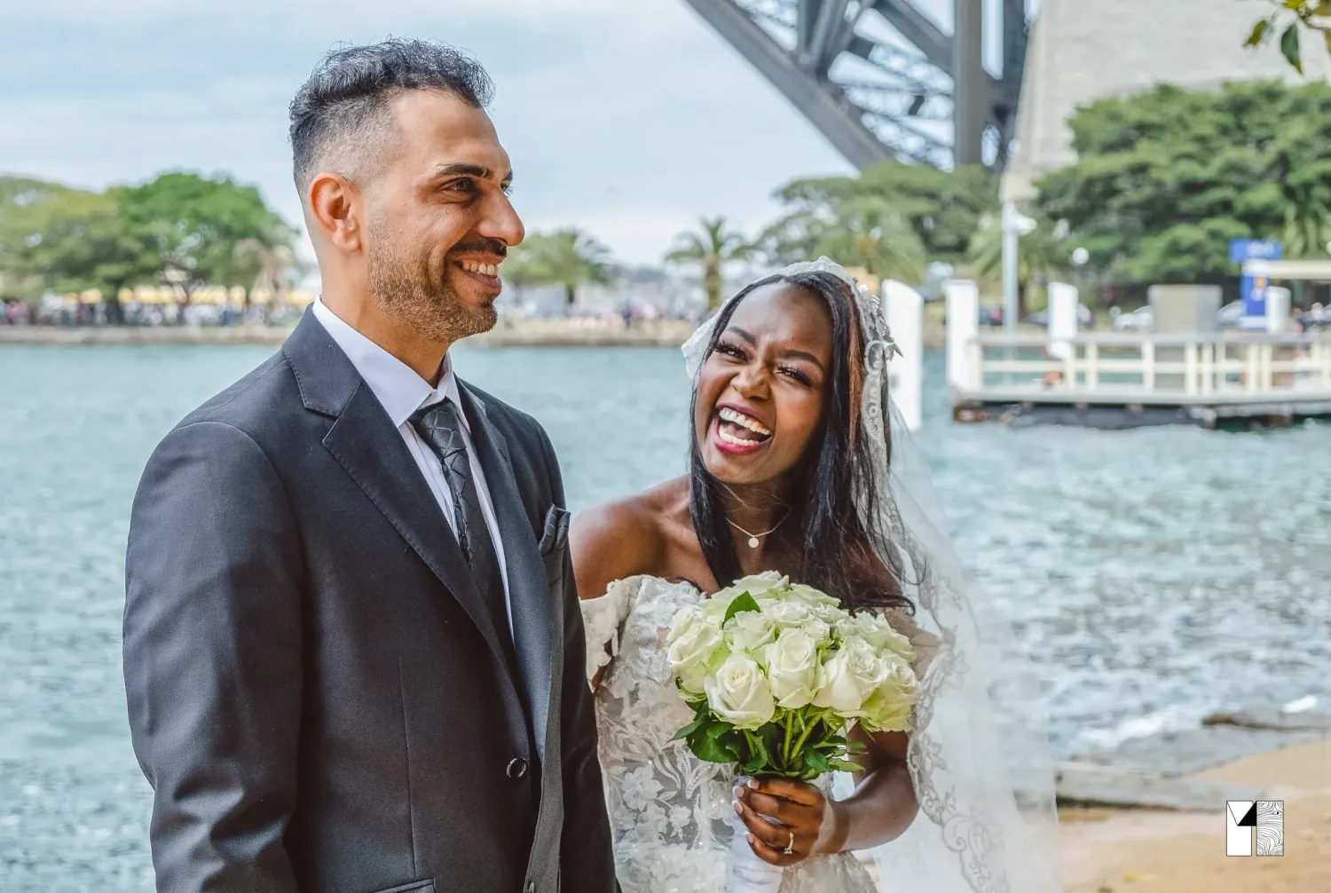 Smiling interracial couple on wedding day with white rose bouquet near harbor bridge.