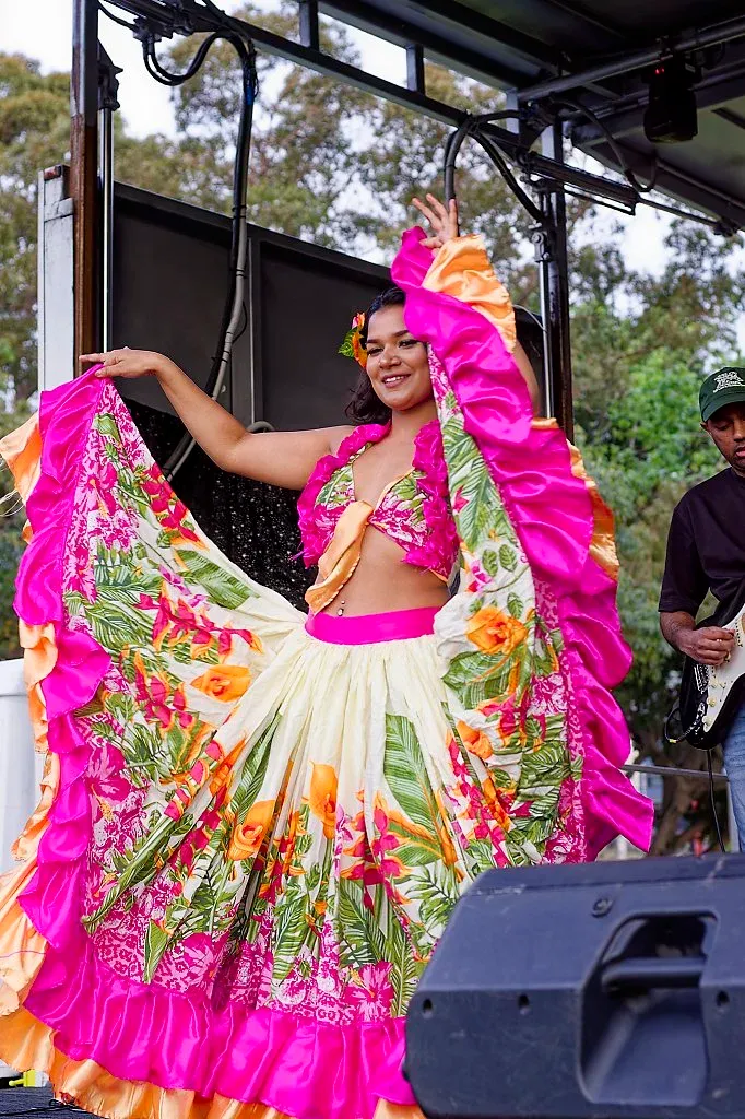 Woman in vibrant pink and floral costume performing a traditional dance on stage.