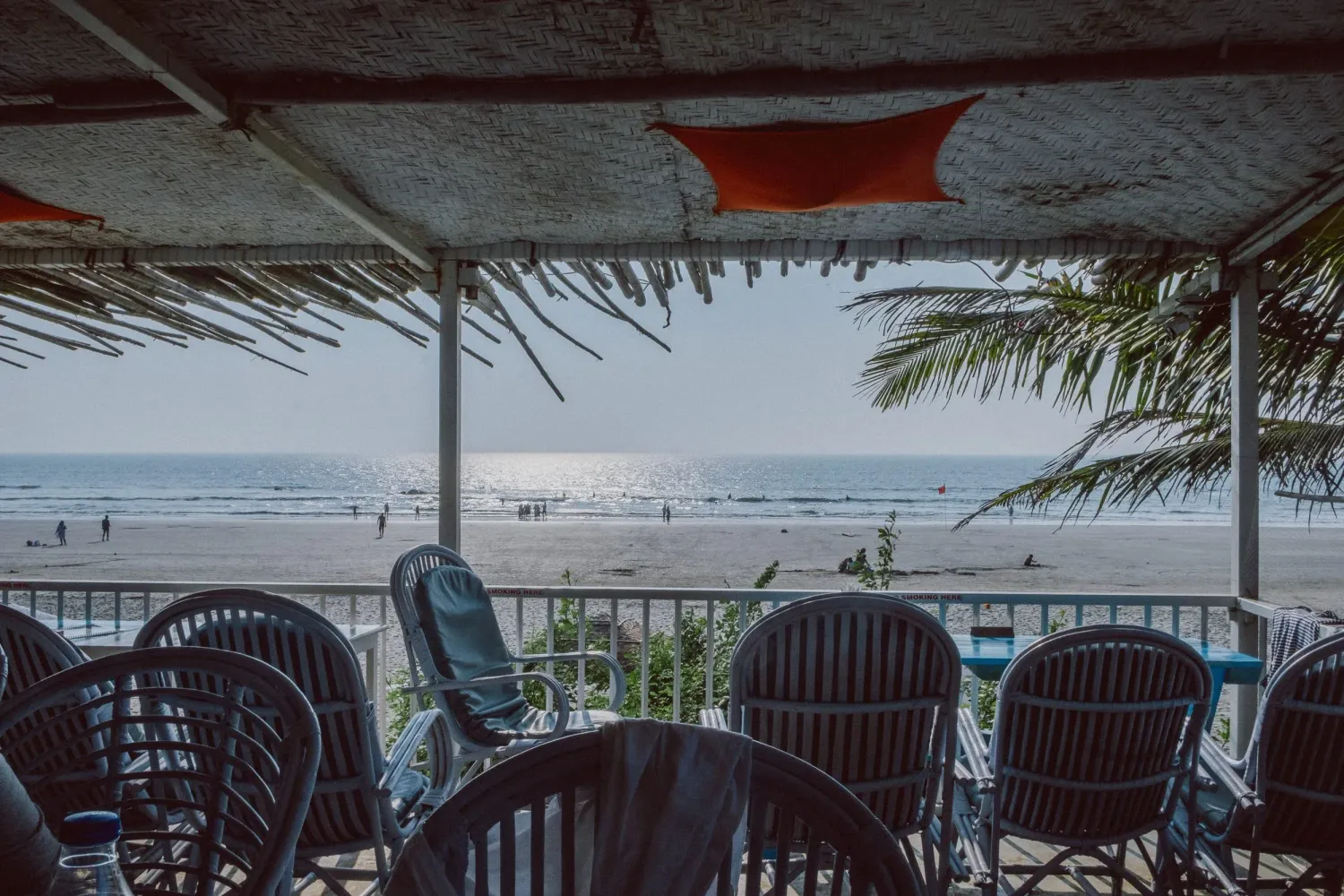 Beachfront restaurant terrace with wicker chairs overlooking sunny sandy beach and ocean view.