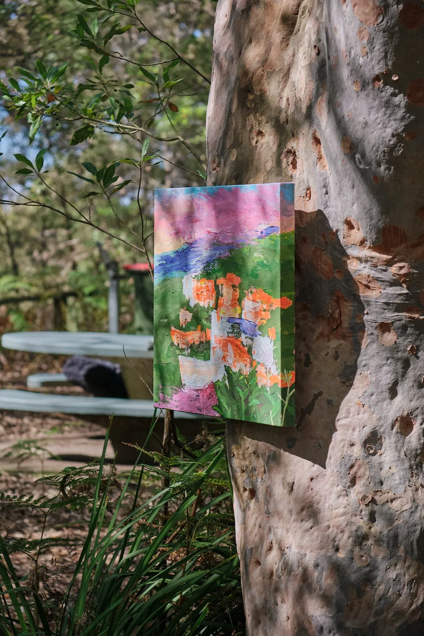 Colorful landscape painting with orange flowers displayed against a eucalyptus tree outdoors.