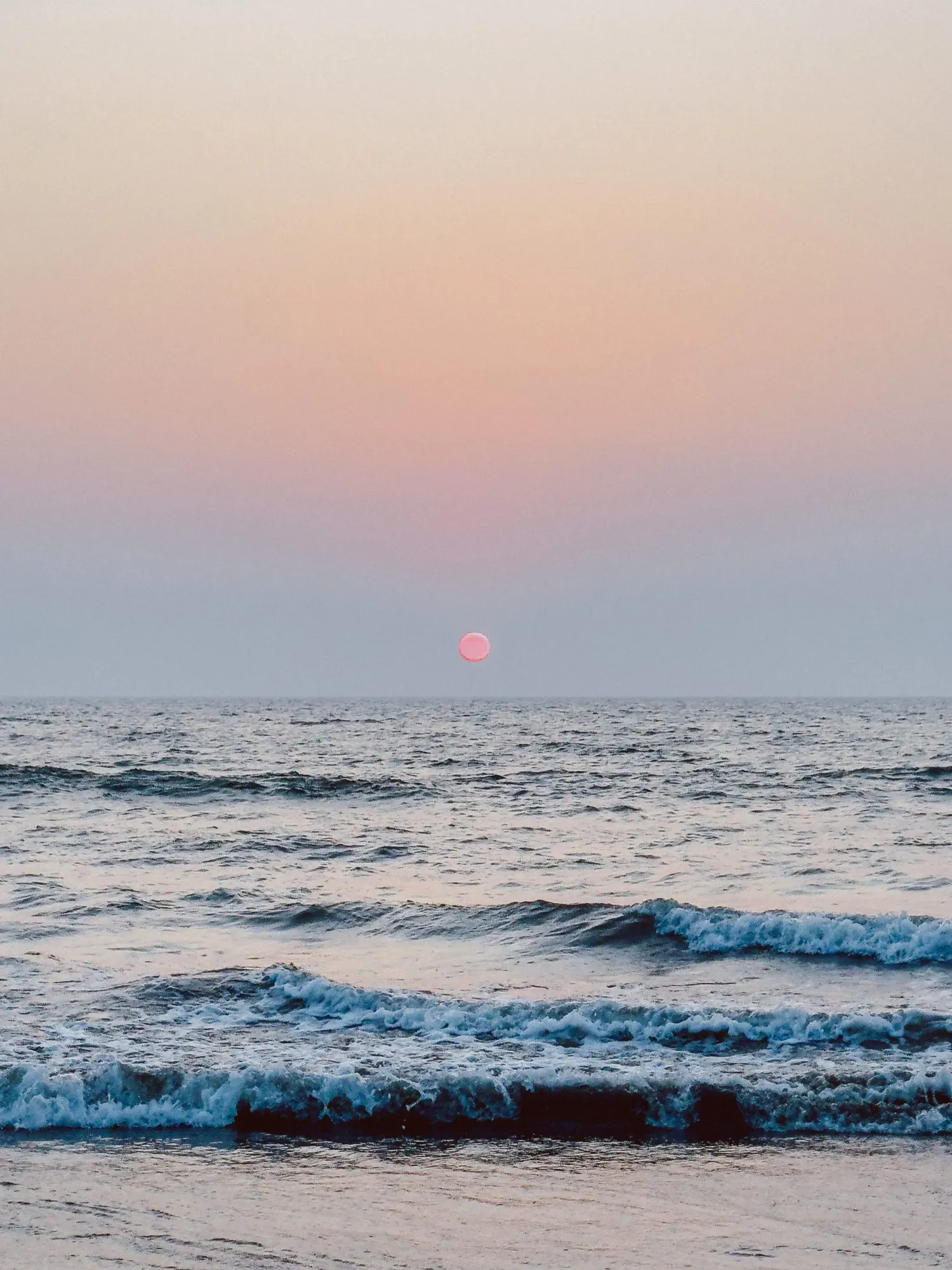 Pink and red sunset sun setting over ocean horizon with rolling waves at dusk.