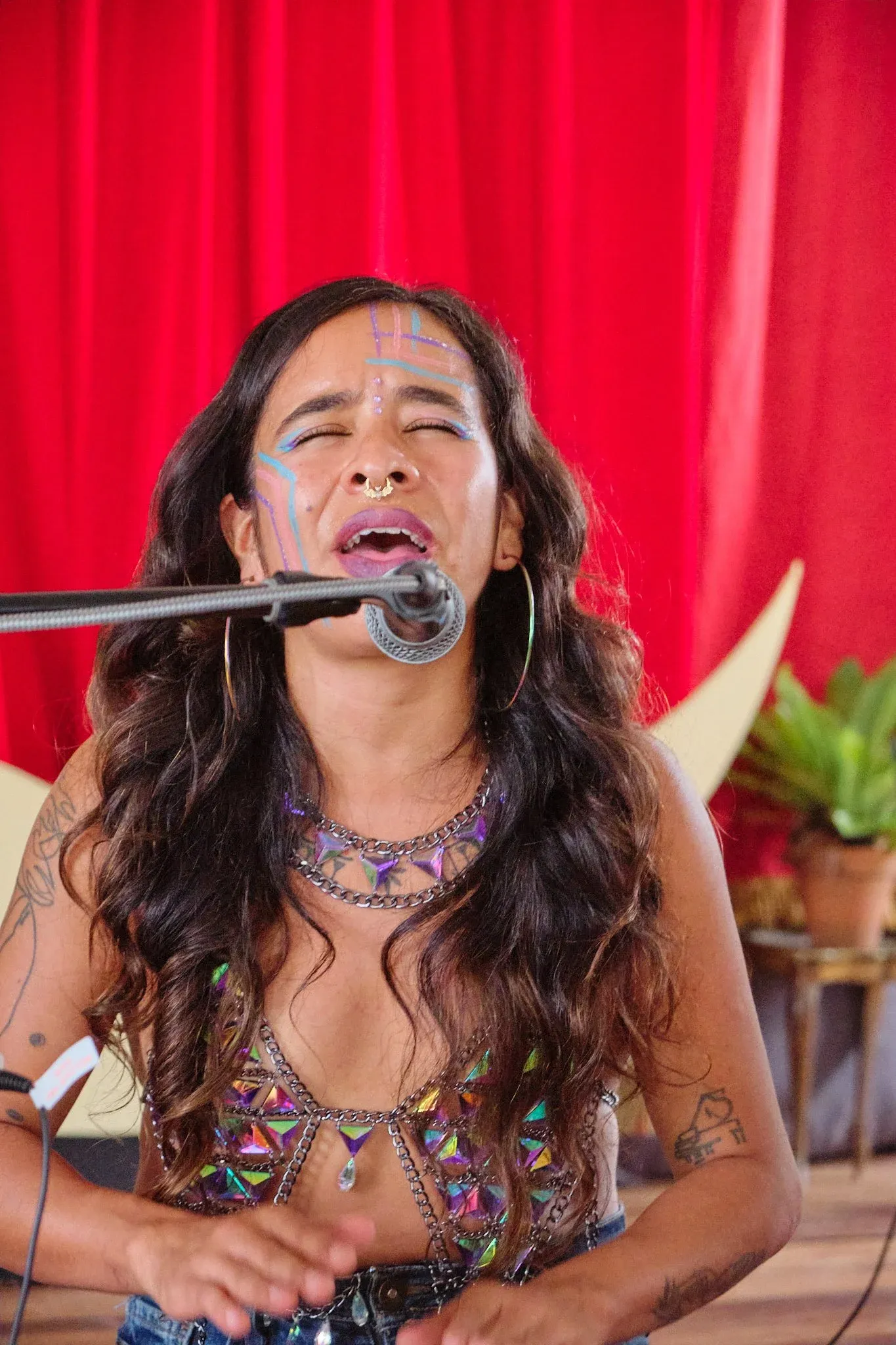 Female vocalist singing intensely at microphone wearing jeweled top with colorful face paint and tattoos.