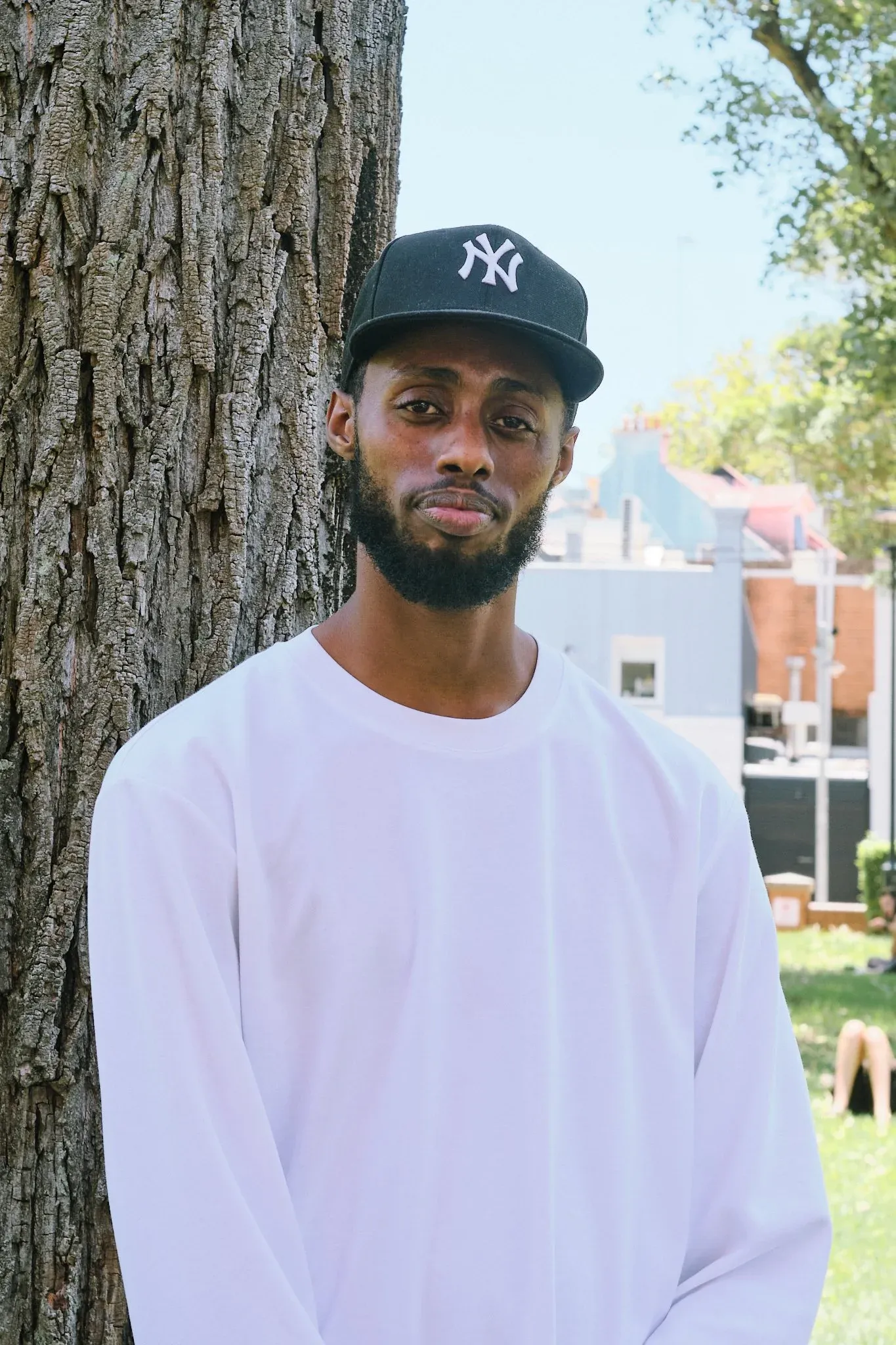 Young Black man with beard wearing white long sleeve shirt and NY Yankees cap leaning against tree outdoors.