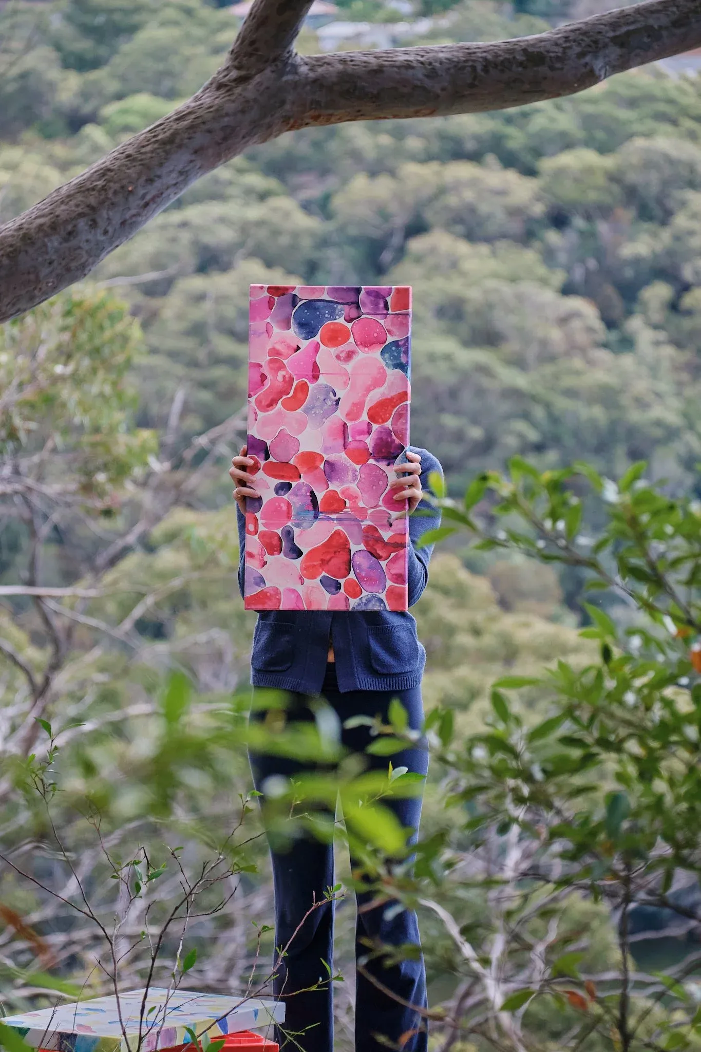Person holding vibrant pink abstract painting up against a lush green forest background.