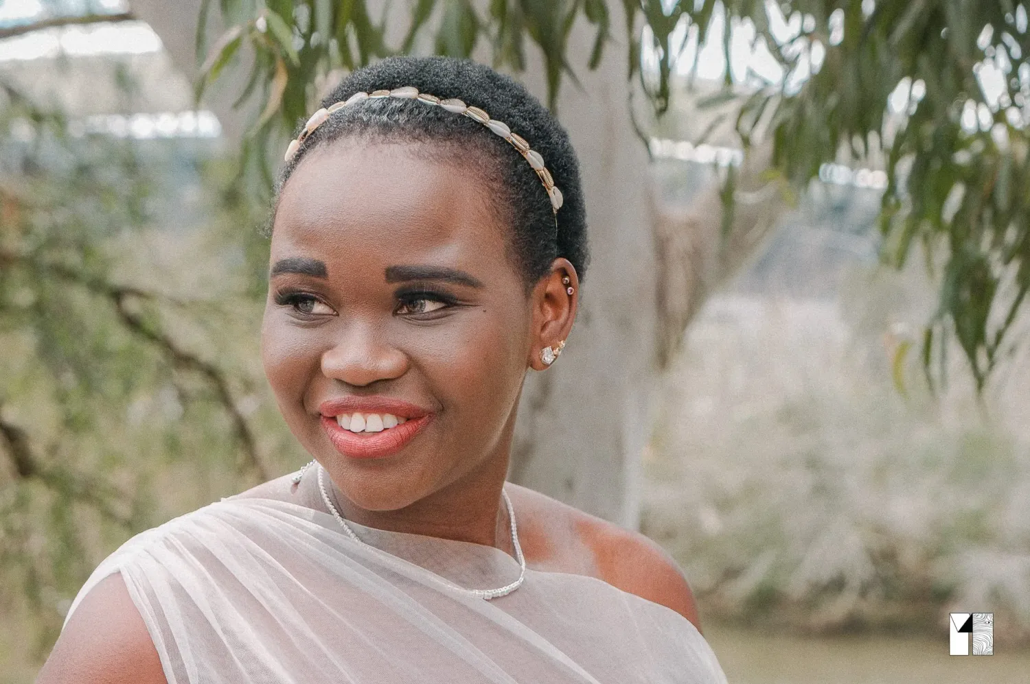 Smiling African bride in sheer wedding dress with pearl headband and earrings among green trees.