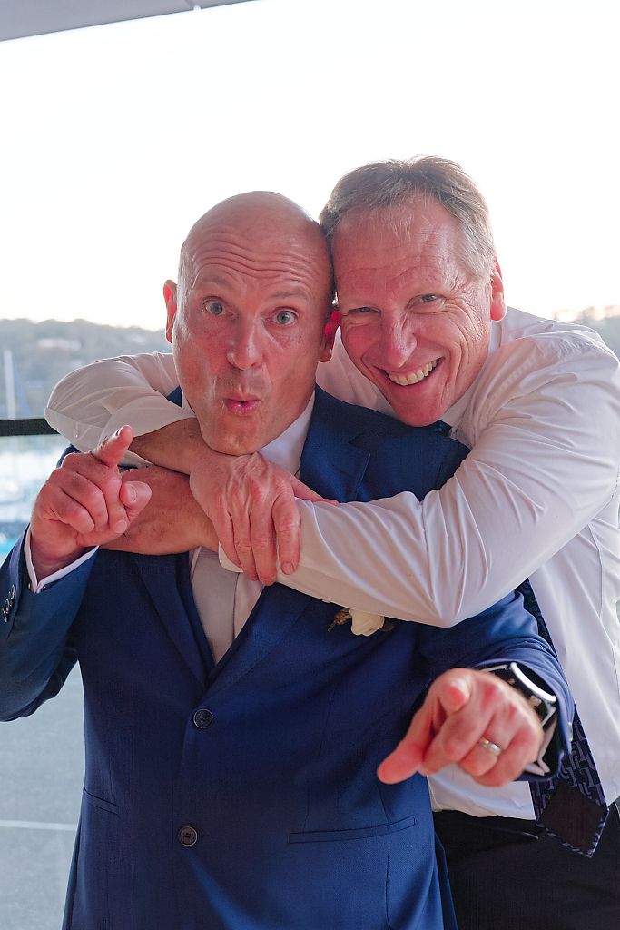 Two cheerful men in suits playfully pose at a wedding, one hugging the other from behind while both point at the camera.