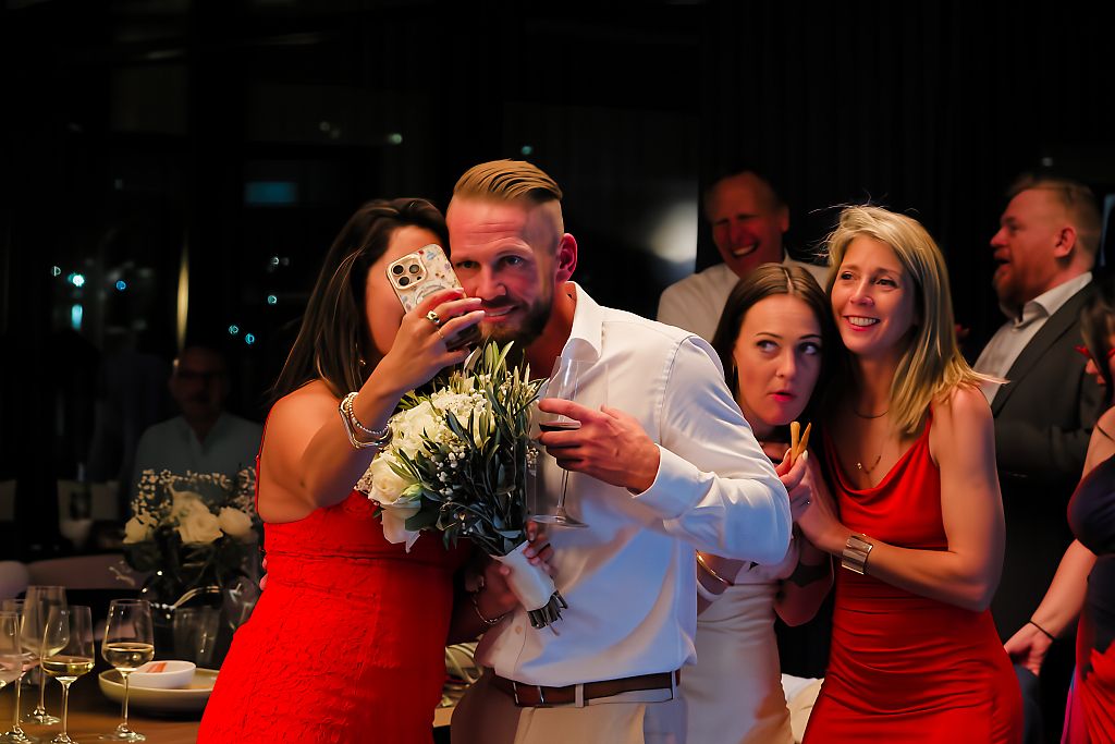 Smiling man in a white shirt holds a white bouquet while a woman photographs him and friends in red dresses celebrate at an indoor party.