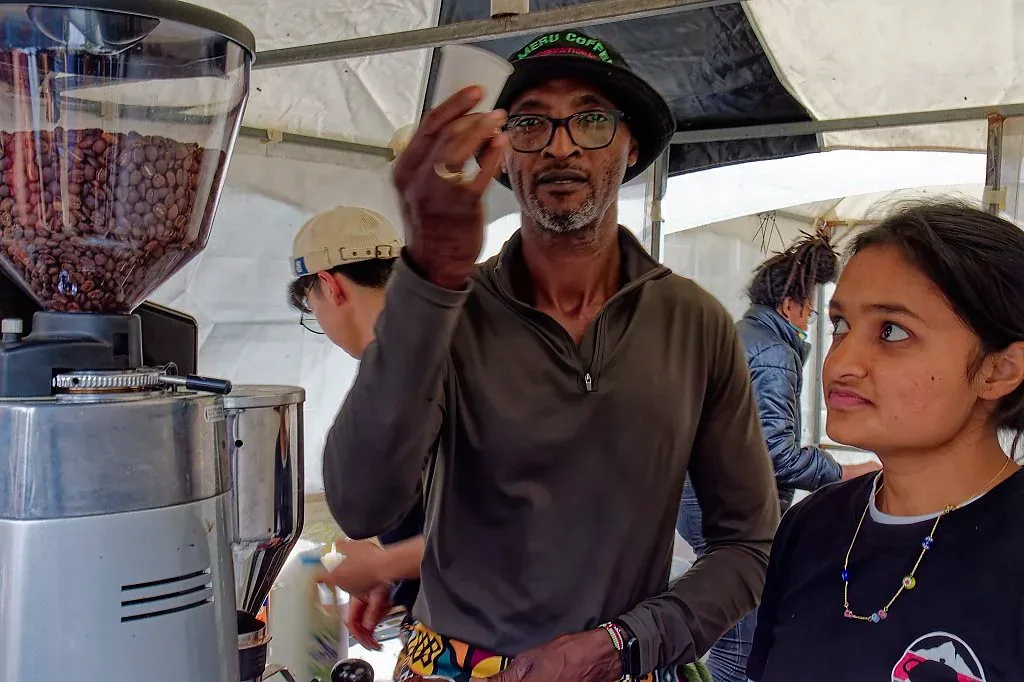 Man explaining coffee grinding process to young woman at specialty coffee stand under tent.