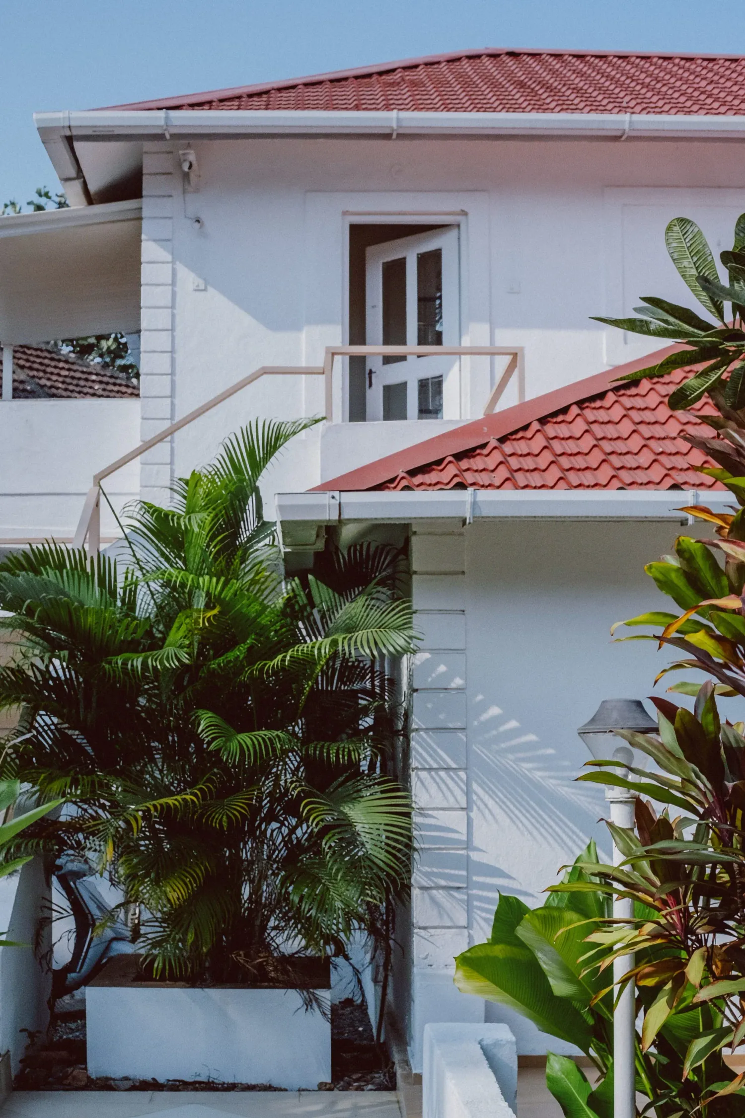 White two-story tropical villa with red tile roof, balcony, and lush palm trees.