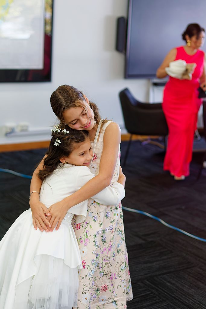 Two young girls in floral and white dresses share a warm hug at a wedding celebration while a woman in a pink dress stands nearby.