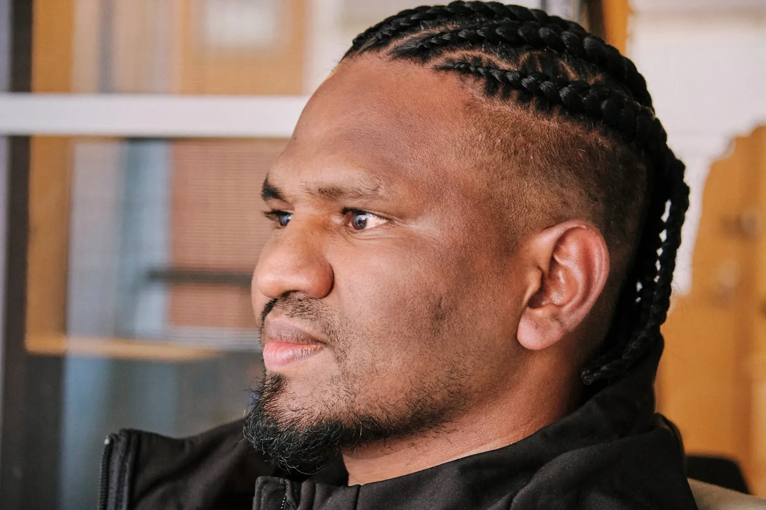 Side profile of man with cornrow braids and beard wearing black jacket in indoor setting.