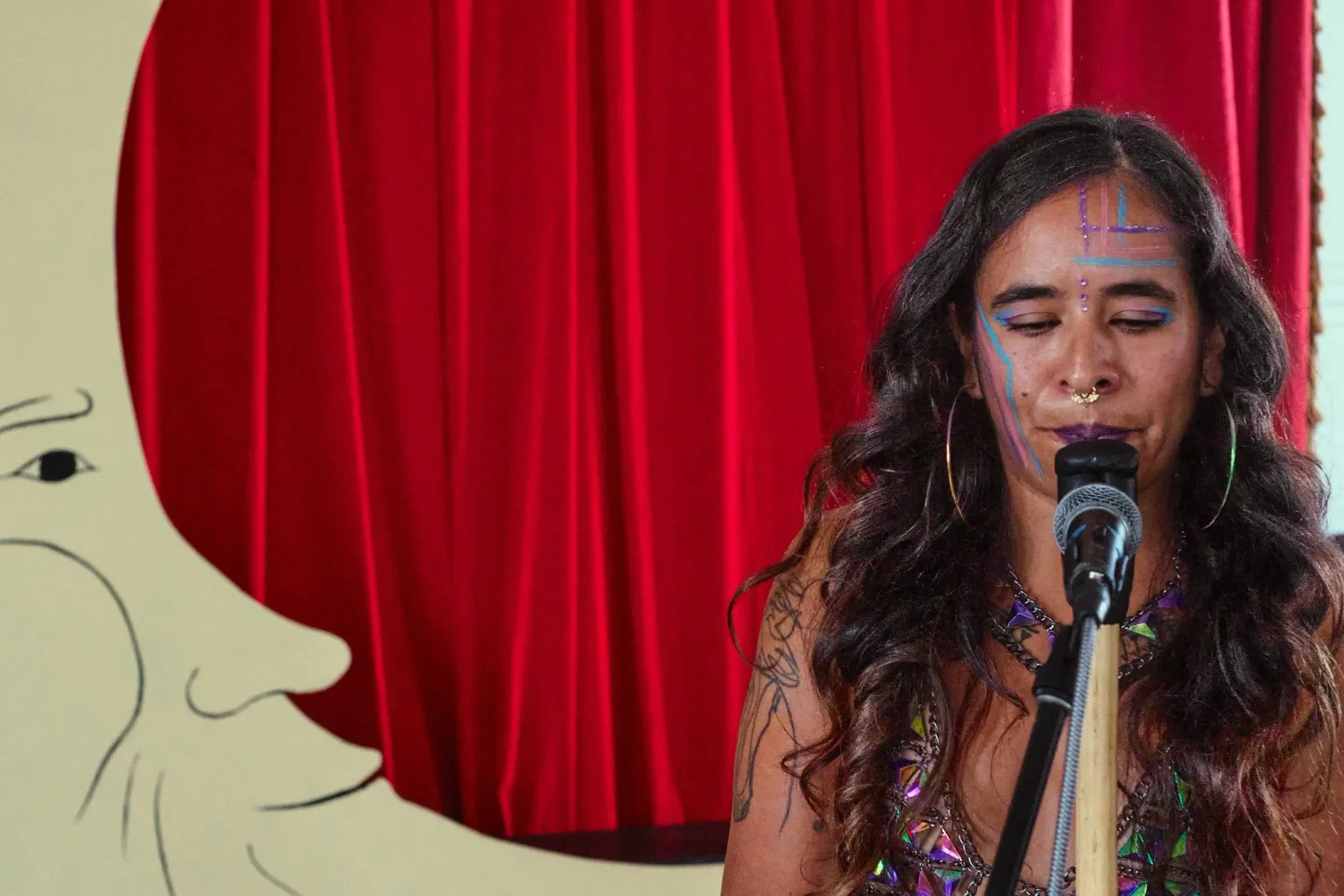 Woman with colorful face paint sings into a microphone in front of a red curtain backdrop.