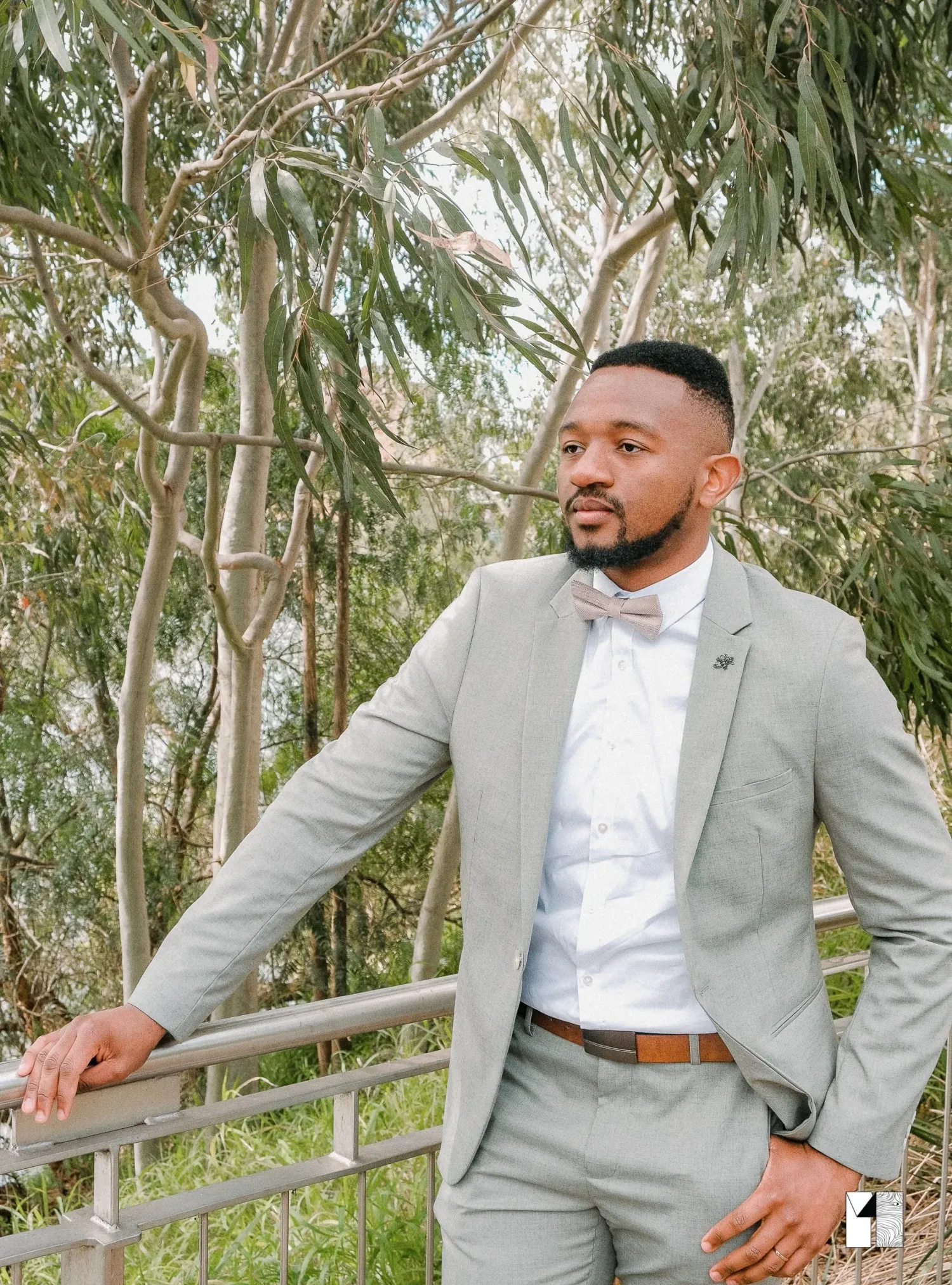 Stylish groom in a grey suit and bow tie posing outdoors near a railing with trees behind him.
