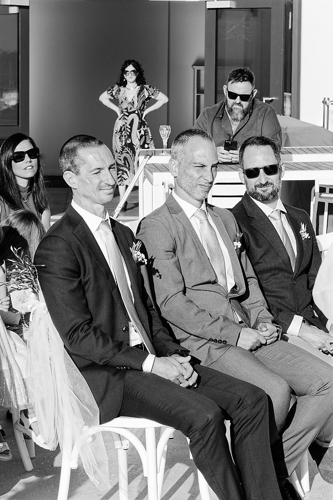Three smiling men in suits and boutonnieres sit side by side among guests in a black and white wedding ceremony photo.