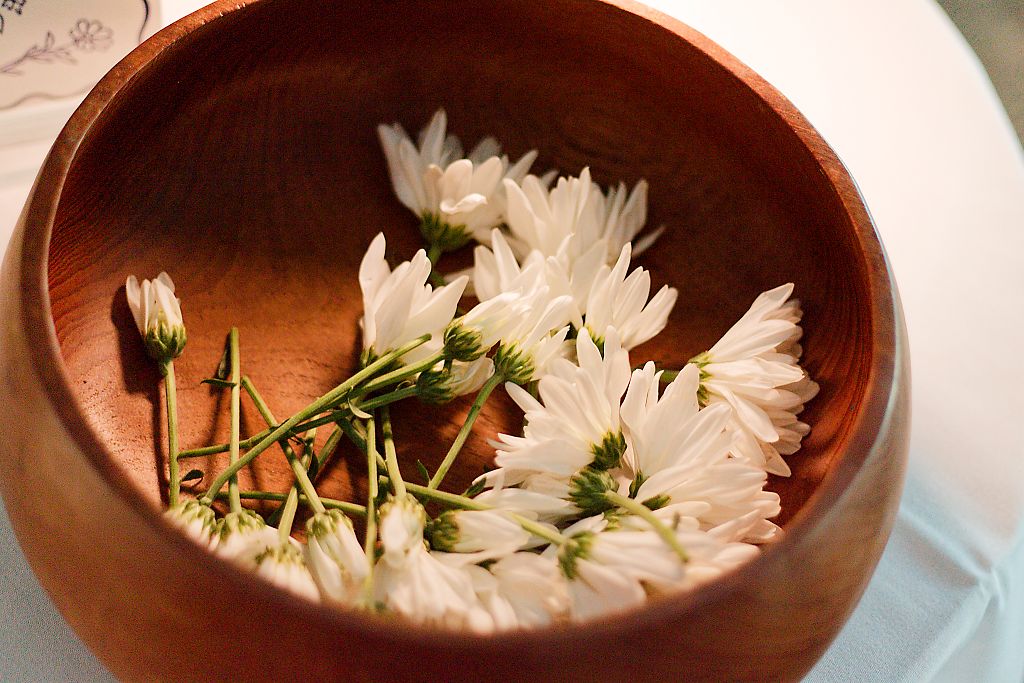 Fresh white chrysanthemum blossoms with green stems resting in a rustic round wooden bowl on a white table.