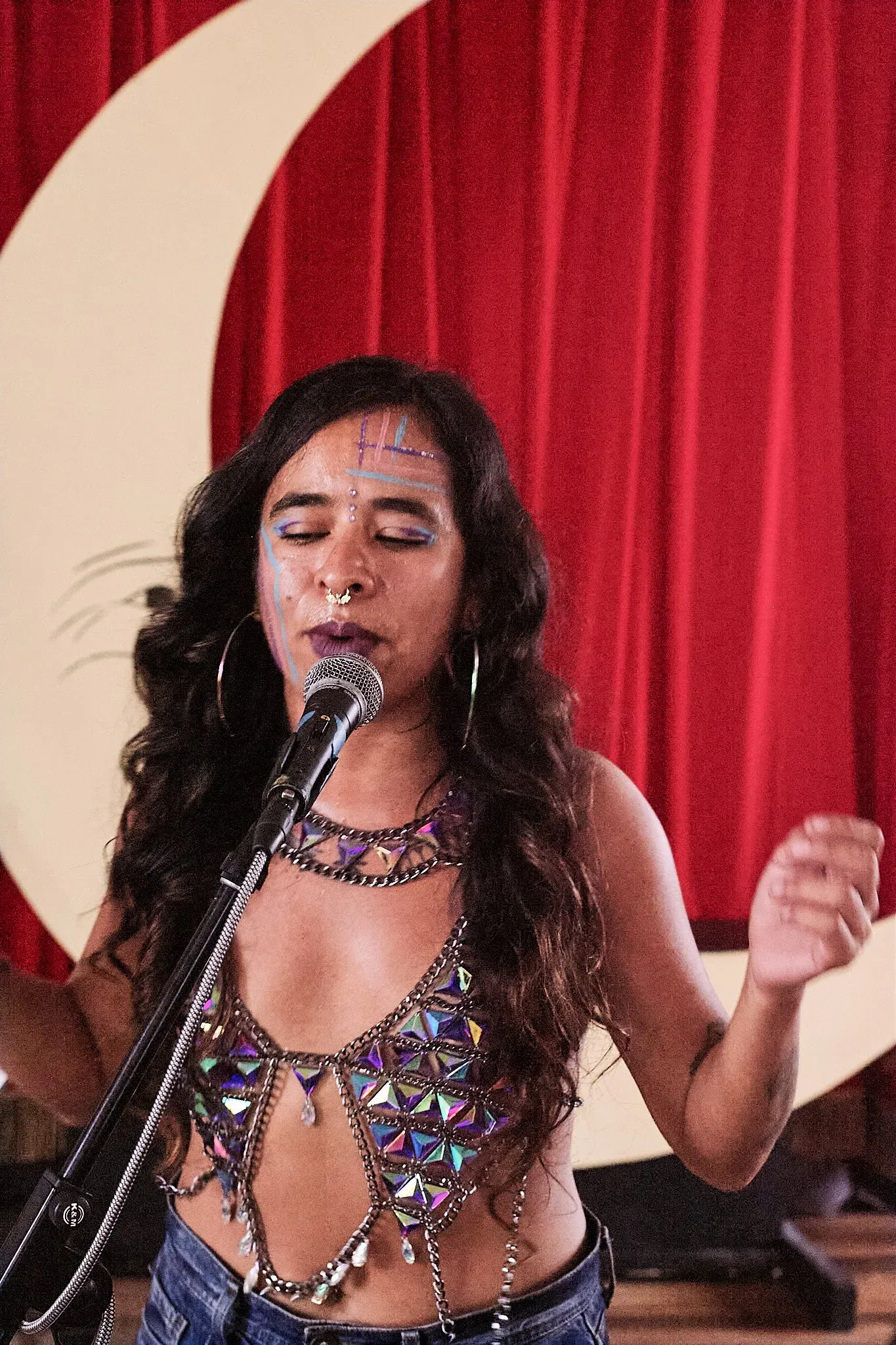 Young woman with face paint singing passionately into microphone wearing sequined top on stage.