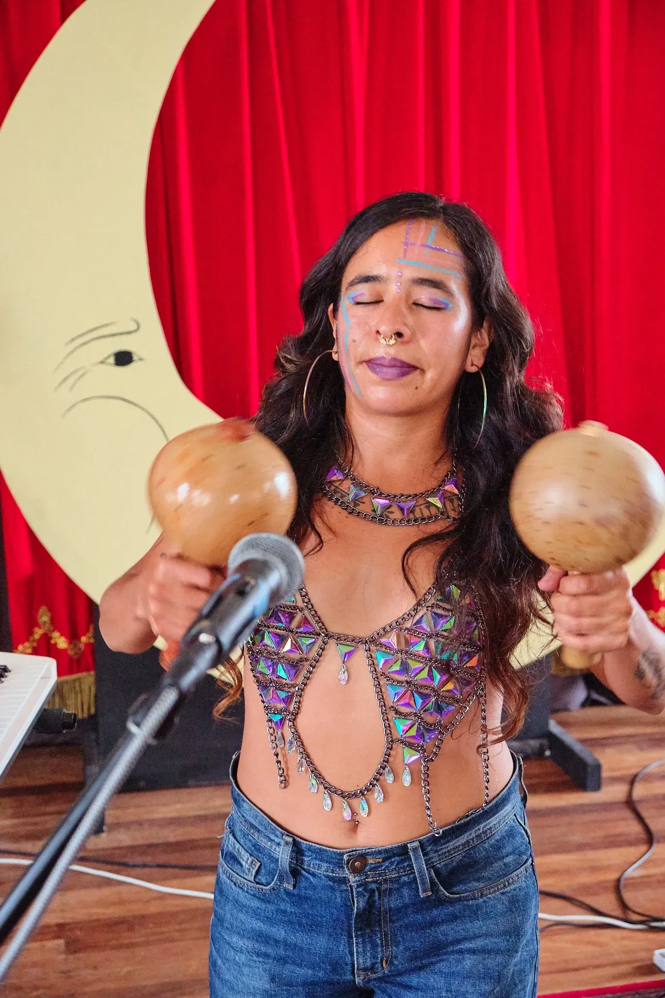 Woman with face paint plays maracas and sings at a microphone in front of a red curtain.