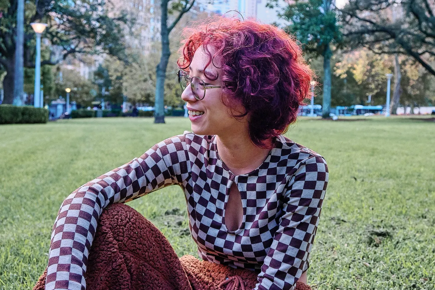 Smiling woman with red curly hair in checkered top sitting on grass in sunny park.