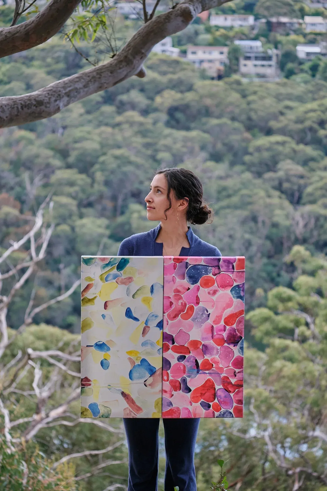 Artist holding two colorful abstract paintings outdoors with Australian bushland in the background.